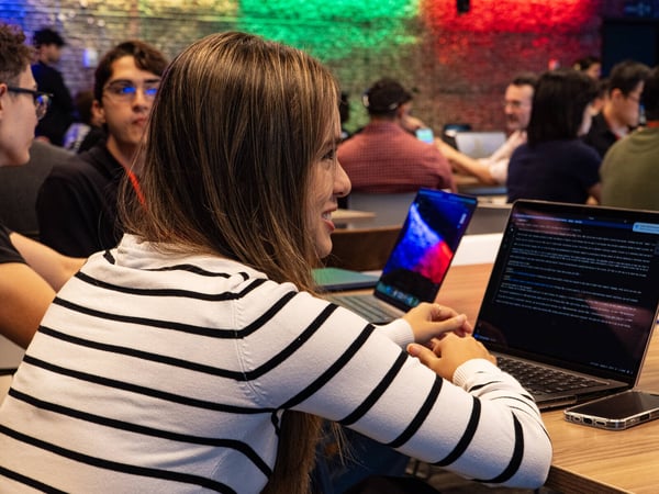 A young woman in a black and white striped sweater sits at a wooden table, smiling while looking at a laptop screen in a crowded, dimly lit room with colorful background lighting.