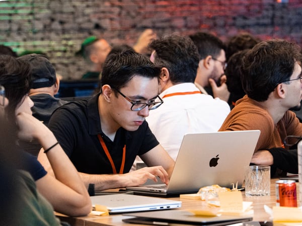 A focused young man with glasses works on his laptop at a crowded table during what appears to be a tech event or hackathon.