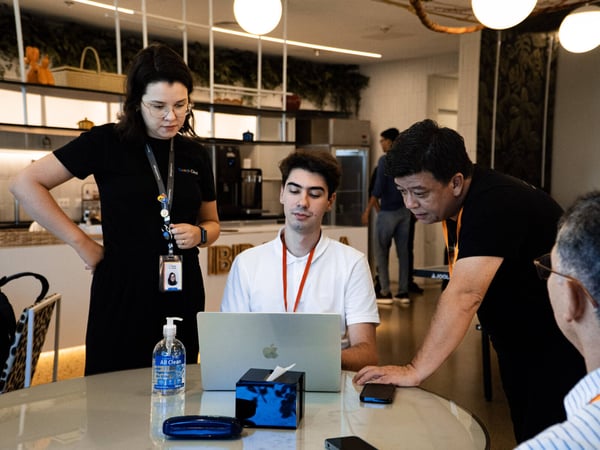 A group of colleagues are gathered around a laptop in a modern office or cafe setting, engaged in a collaborative discussion.