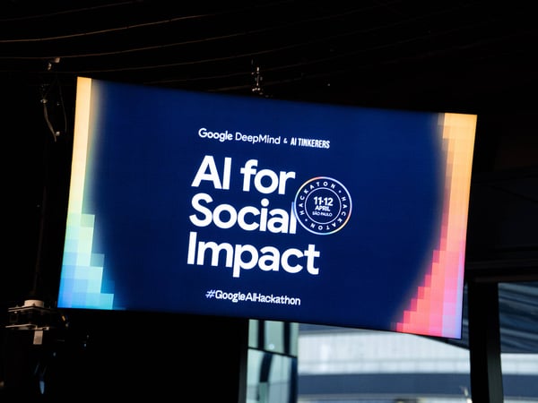 A photograph of a digital display screen announcing an 'AI for Social Impact' hackathon organized by Google DeepMind and AI Tinkerers.