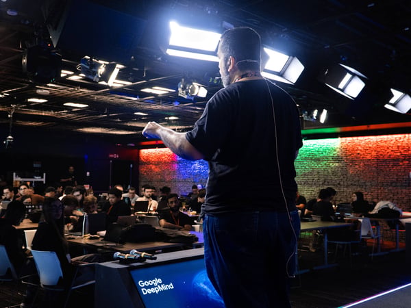 A man stands on a stage with his back to the camera, addressing an audience seated at tables with laptops in a studio-like setting with bright overhead lights.