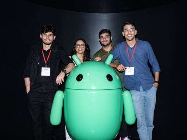 Four young adults are smiling and posing for a photo with a large, glossy green Android mascot statue against a solid black background.