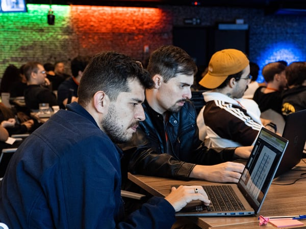 Two men are intently focused on a laptop screen in a dimly lit indoor space with a brick wall background illuminated by green and red lights.