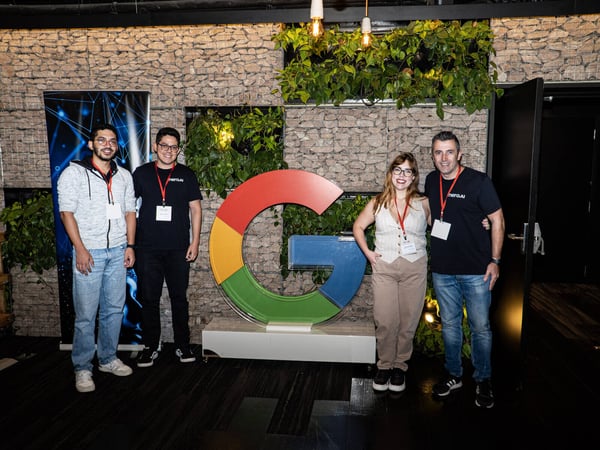 A group of four people, three men and one woman, posing for a photo in front of a large, colorful Google 'G' logo in an indoor setting with a stone wall and plants.