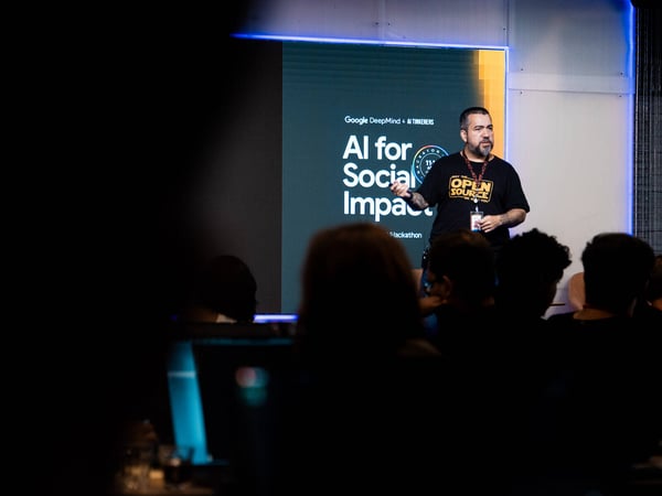 A man stands on a stage giving a presentation titled 'AI for Social Impact' at a Google DeepMind and AI Tinkerers hackathon event.