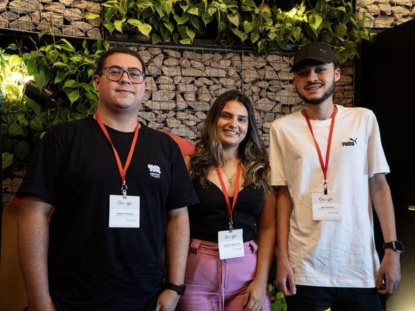 Three young adults, two men and one woman, are posing for a photo while wearing Google-branded lanyards and ID badges in an indoor setting with a stone wall and green plants.