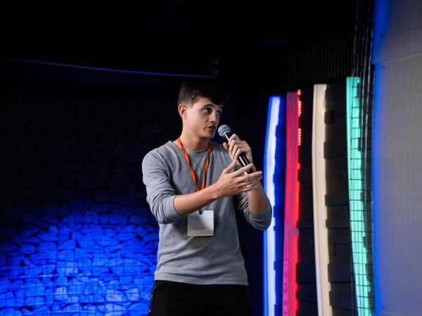 A young man stands on a stage, speaking into a microphone during an event. He is wearing a grey long-sleeved shirt and an orange lanyard with a blank badge.