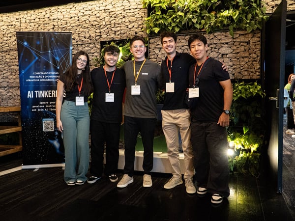 A group of five young adults posing for a photo at an event, standing in front of a stone wall decorated with green plants.