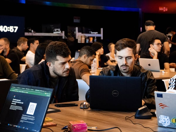 A group of people are focused on their laptops in a dimly lit indoor space, likely a hackathon or collaborative workspace.