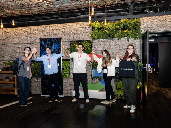 A group of five young adults pose for a photo in an indoor event space, standing in front of a stone wall decorated with plants and a large Google logo.