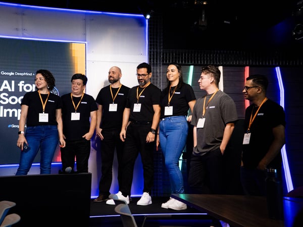 A group of seven people stands on a stage during a Google DeepMind event, with a screen in the background displaying 'AI for Social Impact'.