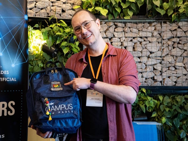 A man smiling and pointing at a 'Google AI Builders' patch on a blue Google Campus São Paulo backpack.