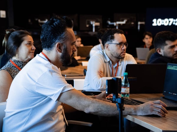 A group of people are seated at desks in a dimly lit room, attending a workshop or presentation. A man in the foreground is working on a laptop with a camera mounted on a tripod nearby.