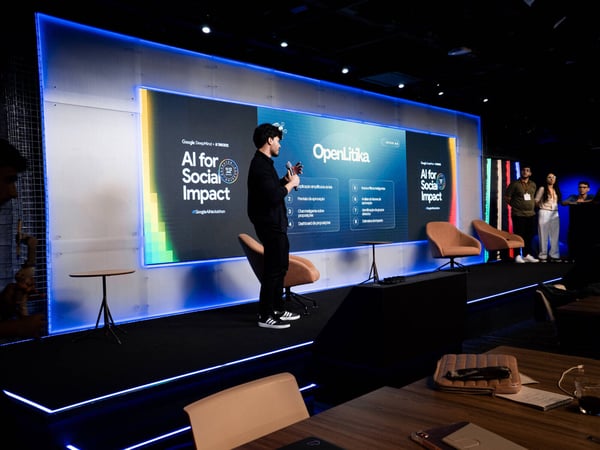 A man is giving a presentation on a stage in front of a large digital screen displaying information about 'AI for Social Impact' and 'OpenLitika'.