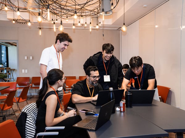 A group of young adults are collaborating and smiling while working on laptops in a modern office space featuring a unique, tangled light fixture.
