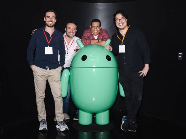 Four men are smiling and posing for a group photo with a large, glossy green Android mascot statue against a black backdrop.