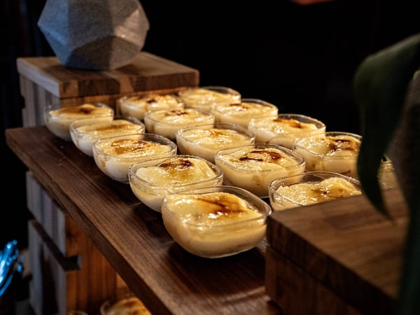 A photograph showing multiple small glass bowls filled with a creamy dessert, likely crème brûlée, arranged neatly on a wooden tiered display stand.