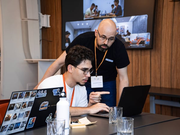 Two men are focused on a laptop screen in a modern office or event space, with one man pointing at the display while the other types.