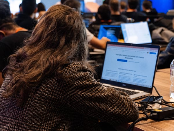 A person with long, wavy brown hair is seen from behind, sitting at a desk and using a laptop in a crowded room filled with other people working on computers.