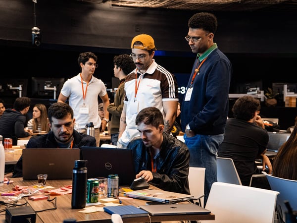 A group of young people are gathered around a table with laptops in a dimly lit indoor setting, appearing to be collaborating or debugging code.