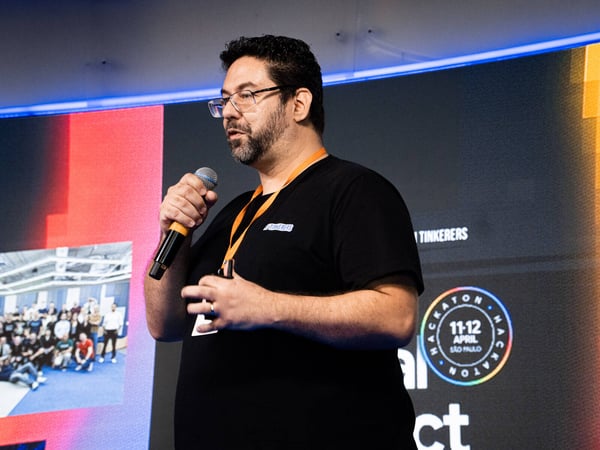 A man with glasses and a beard is speaking into a microphone during a presentation at an event, with a large screen displaying hackathon details behind him.