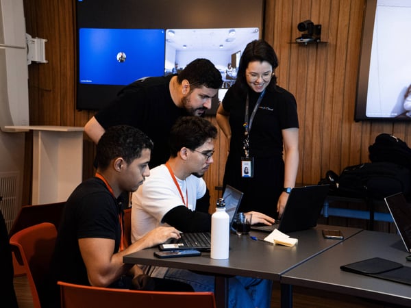 A group of four individuals are gathered around a table with laptops, appearing to be engaged in a collaborative technical discussion or debugging session in a modern office setting.