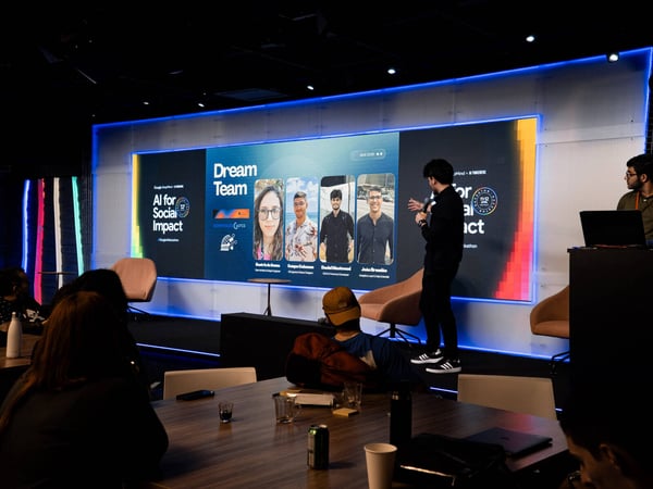 A man stands on a stage giving a presentation in front of a large screen displaying 'Dream Team' and photos of four individuals at an event titled 'AI for Social Impact'.