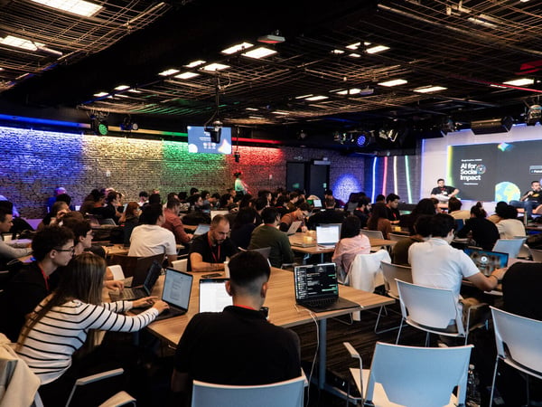 A large group of people attending a tech presentation titled 'AI for Social Impact' in a modern, industrial-style venue with colorful lighting.