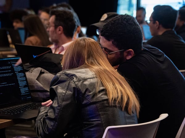 Two individuals are seen from behind, focused on a laptop screen in a crowded, dimly lit room that appears to be a tech workshop or hackathon.