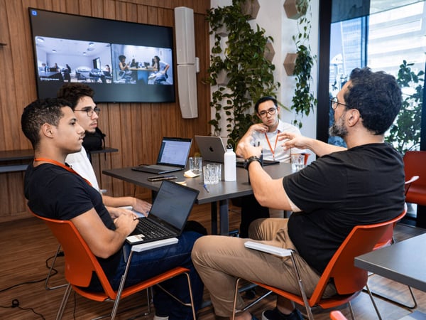 A group of four men are engaged in a collaborative discussion around a table in a modern office, with laptops open and a video conference screen in the background.