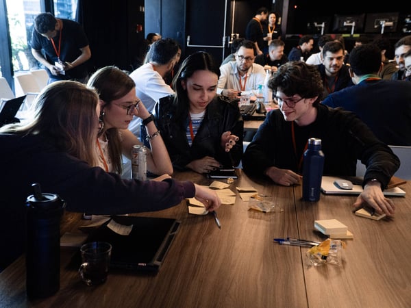 A group of young adults are gathered around a wooden table in a dimly lit indoor space, actively collaborating on a project using sticky notes and laptops.