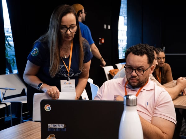 A woman in a blue 'Women in AI' t-shirt leans over to collaborate with a man working on a laptop at a tech event.