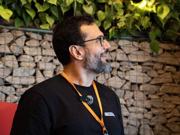 A man with a beard and glasses is looking upwards and to the side, wearing a black t-shirt and an orange lanyard against a stone wall background.
