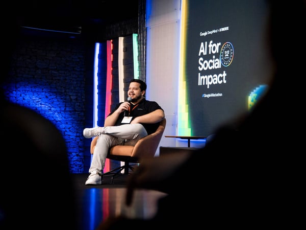 A man sits on a stage holding a microphone, speaking in front of a screen that reads 'AI for Social Impact' during a Google AI Hackathon event.
