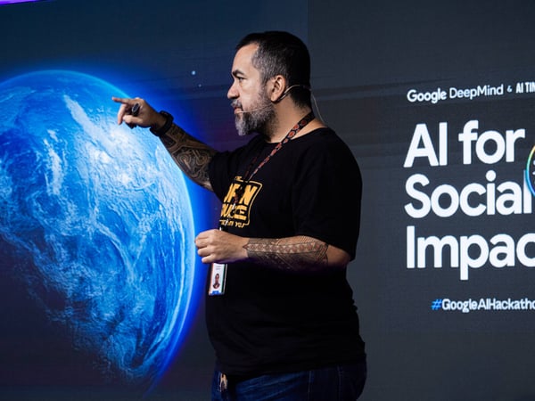 A man with prominent arm tattoos is giving a presentation in front of a large screen displaying an image of Earth and text about AI for social impact.