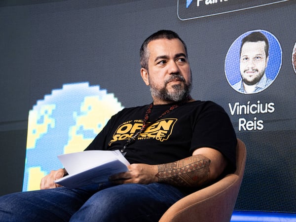 A man with a beard and an arm tattoo sits on a stage during a presentation, with a large digital screen behind him showing a pixelated globe and a speaker profile.