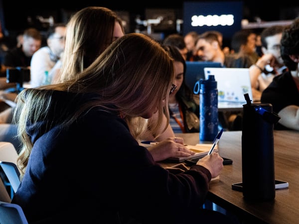 A candid photograph of people participating in an indoor event, with a focus on a young woman in the foreground writing in a notebook.
