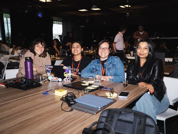 Four women are smiling at the camera while sitting around a wooden table in a busy indoor workspace or event venue.