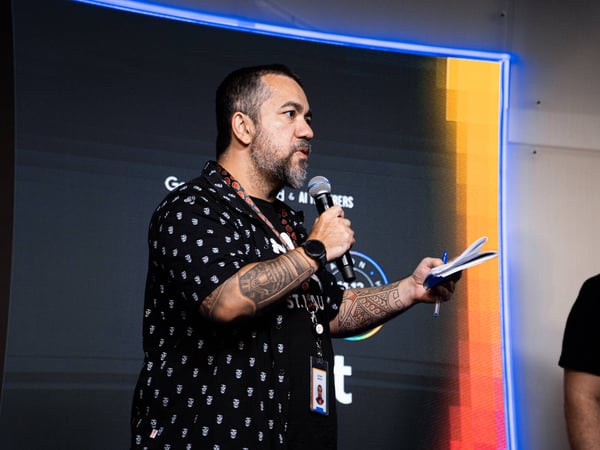 A man with extensive arm tattoos is speaking into a microphone while holding a pen and notepad at a tech event.