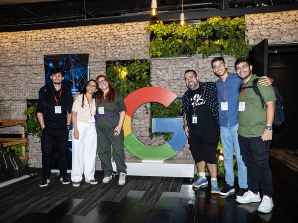 A group of six people posing for a photo in front of a large, colorful Google 'G' logo in an indoor space with a stone wall and plants.