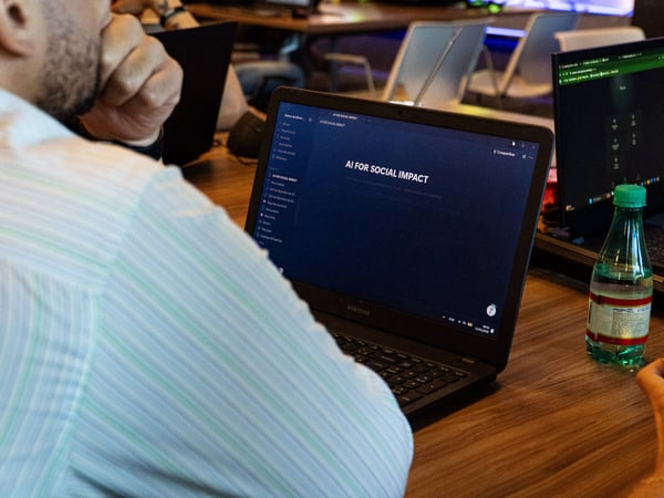 A person is viewed from behind, looking at a laptop screen that displays the text 'AI FOR SOCIAL IMPACT' in a workspace setting.