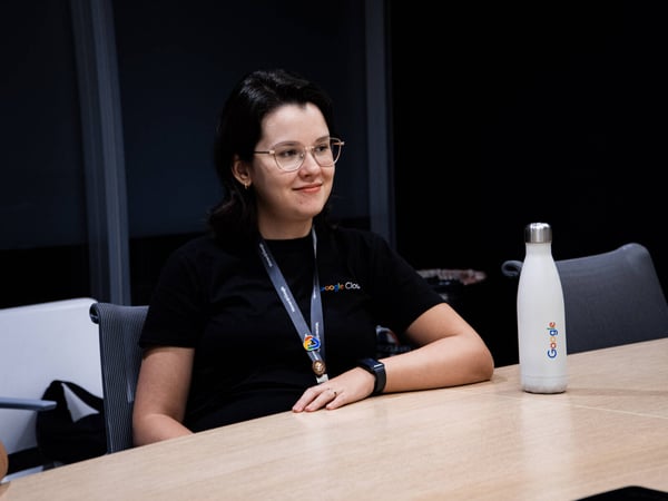 A woman wearing glasses and a Google Cloud t-shirt is sitting at a wooden table in an office setting next to a white Google water bottle.