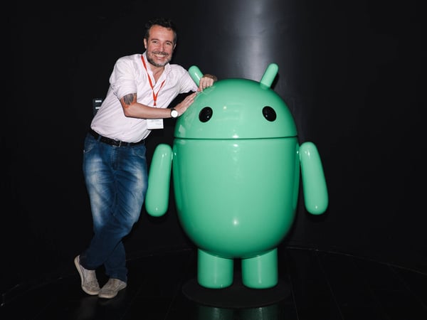A man in a white shirt and jeans is smiling and leaning against a large, green Android mascot statue against a solid black background.