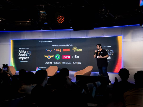 A man stands on a stage giving a presentation in front of a large digital screen displaying various partner logos for an AI event in São Paulo.