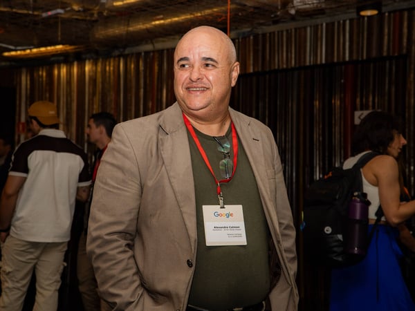 A bald man in a tan blazer and olive green shirt smiles at an indoor event while wearing a Google lanyard and name tag.