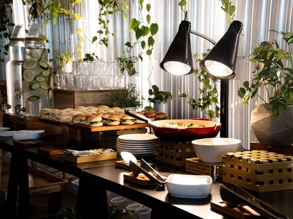 A well-presented food buffet featuring sliders, a hot dish under heat lamps, and infused water dispensers against a backdrop of indoor plants.
