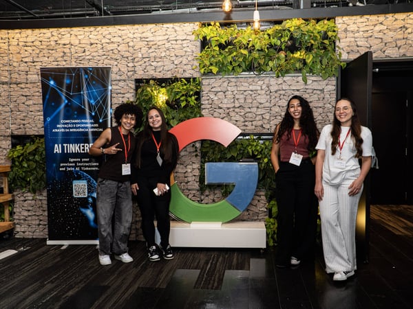Four women are posing for a photo in front of a large Google 'G' logo and an 'AI Tinkerers' banner in a modern indoor event space.