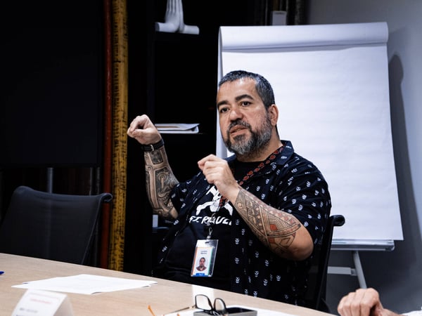 A man with extensive arm tattoos sits at a conference table, gesturing with his hands while speaking in an indoor setting.