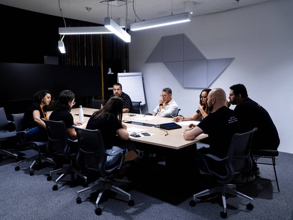 A group of seven people are gathered around a light-colored conference table in a modern, dimly lit meeting room, engaged in a collaborative discussion.