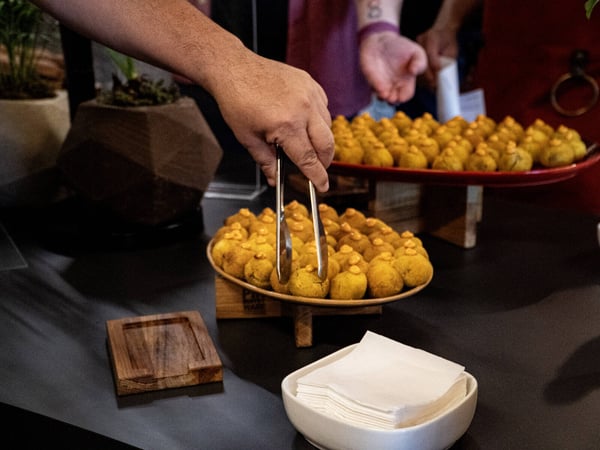 A close-up photograph of a person using metal tongs to select a small, round appetizer from a wooden platter at a social gathering.